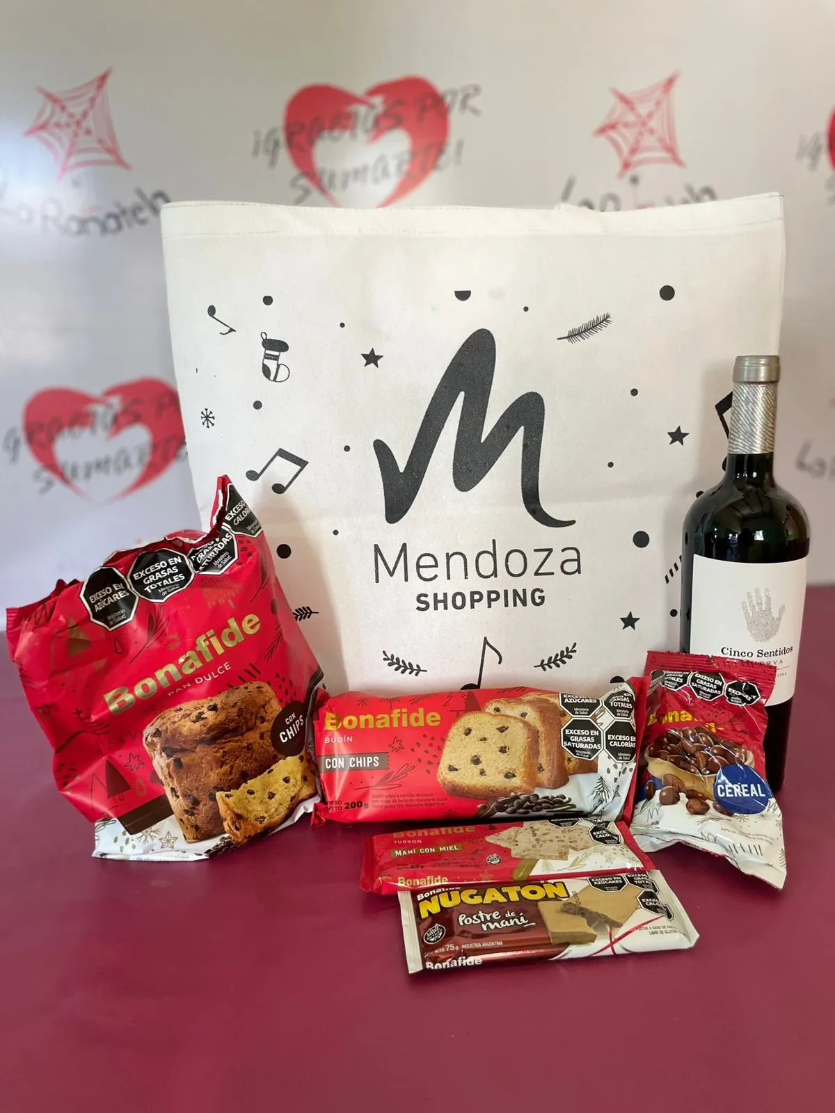 Bolsa de friselina navideña con logo de Mendoza Shopping, junto a pan dulce, budín, turrón, chocolates y una botella de vino