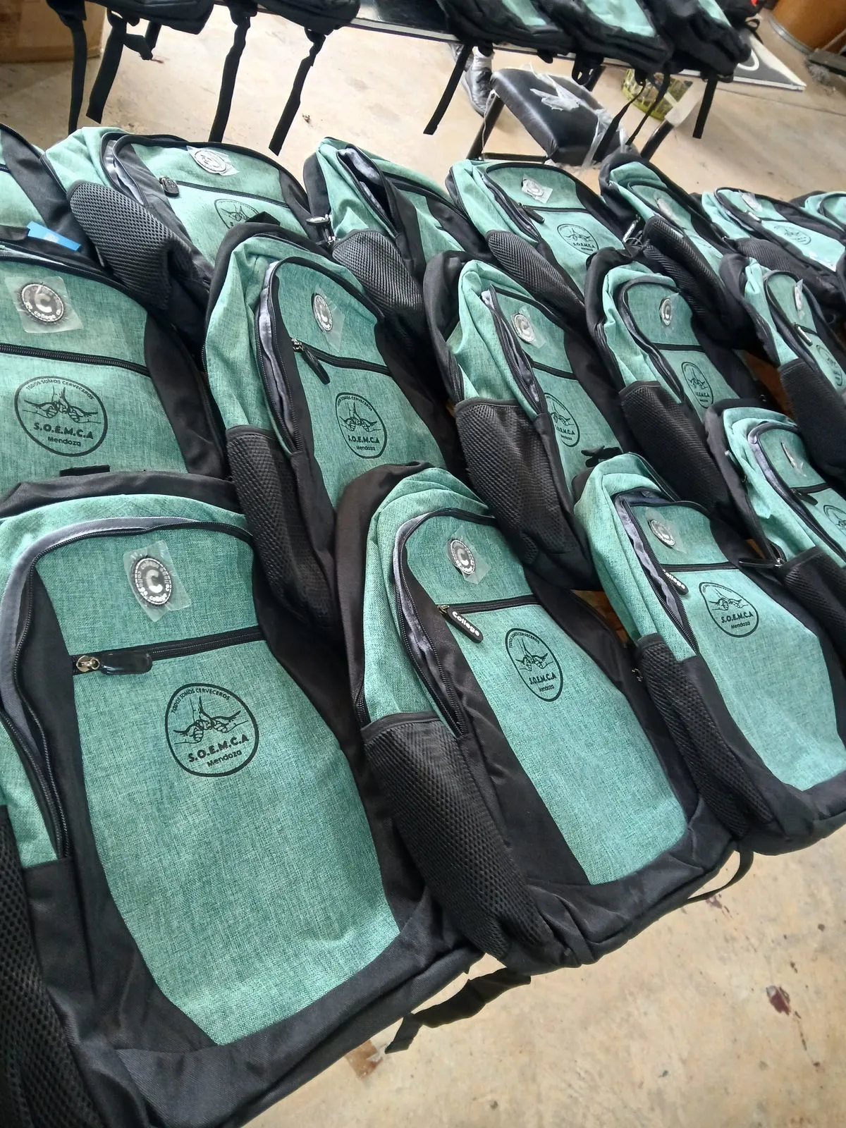 Mochilas verde agua con logo estampado, alineadas sobre una mesa del taller